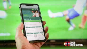 En hånd holder en mobiltelefon med en åpen betting-app foran en TV-skjerm
